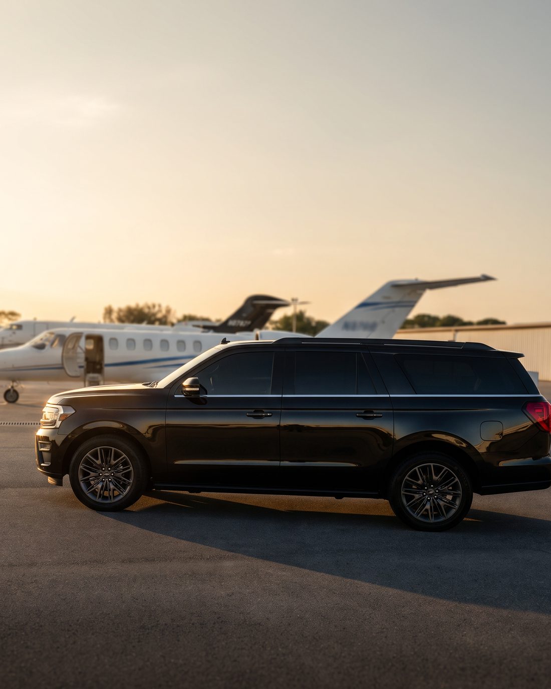A black Ford Expedition Max on a private aviation apron at sunset, a business jet parked behind it